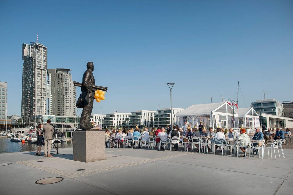 Fotografia z obchodów na ostrodze mola Rybackiego. Na pierwszym planie widoczny pomnik Tadeusza Wendy, a w tle goście zasiadający do stołu.
